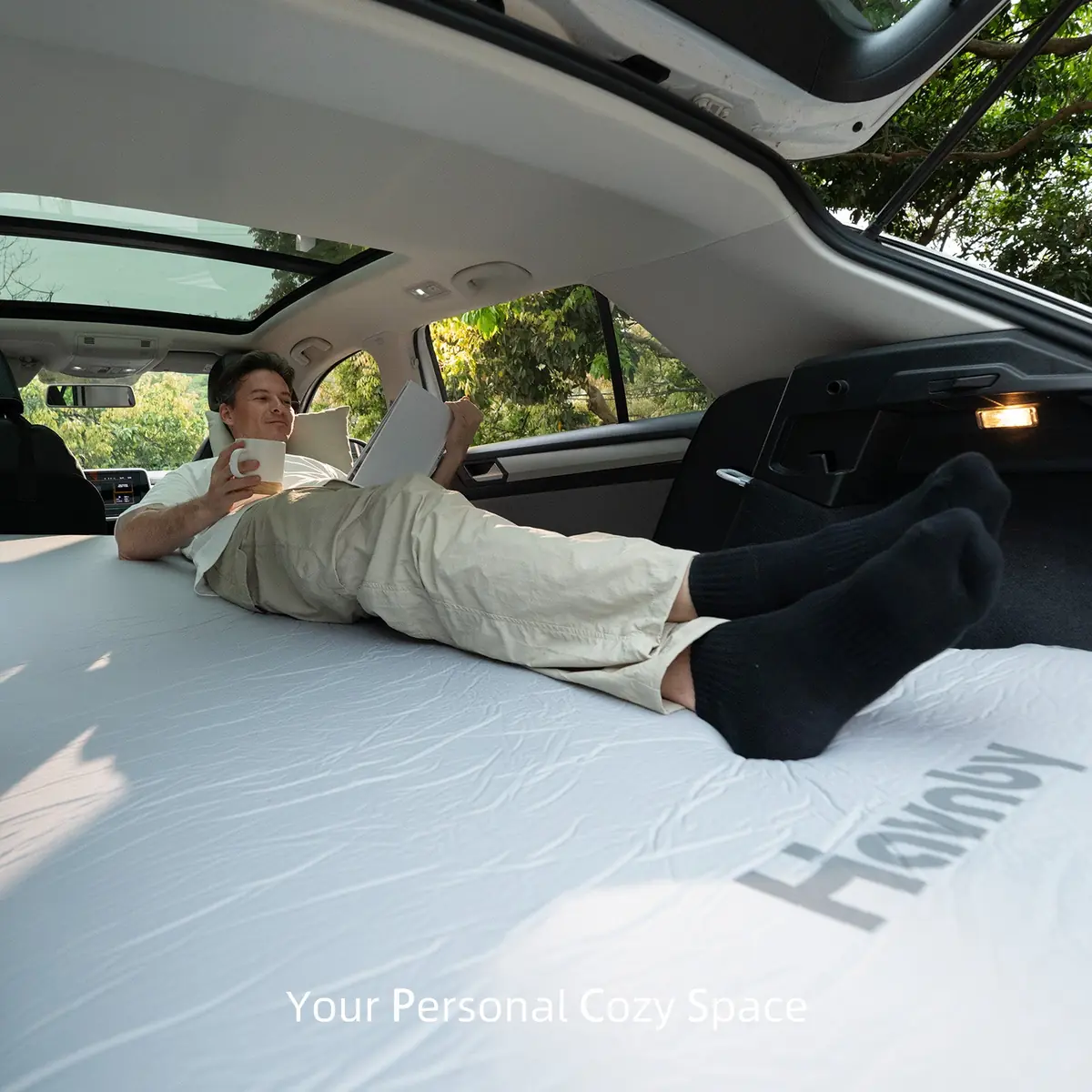 Man reclining on the deployed Havnby CloudCore mattress inside a vehicle cargo area with a laptop and a coffee mug