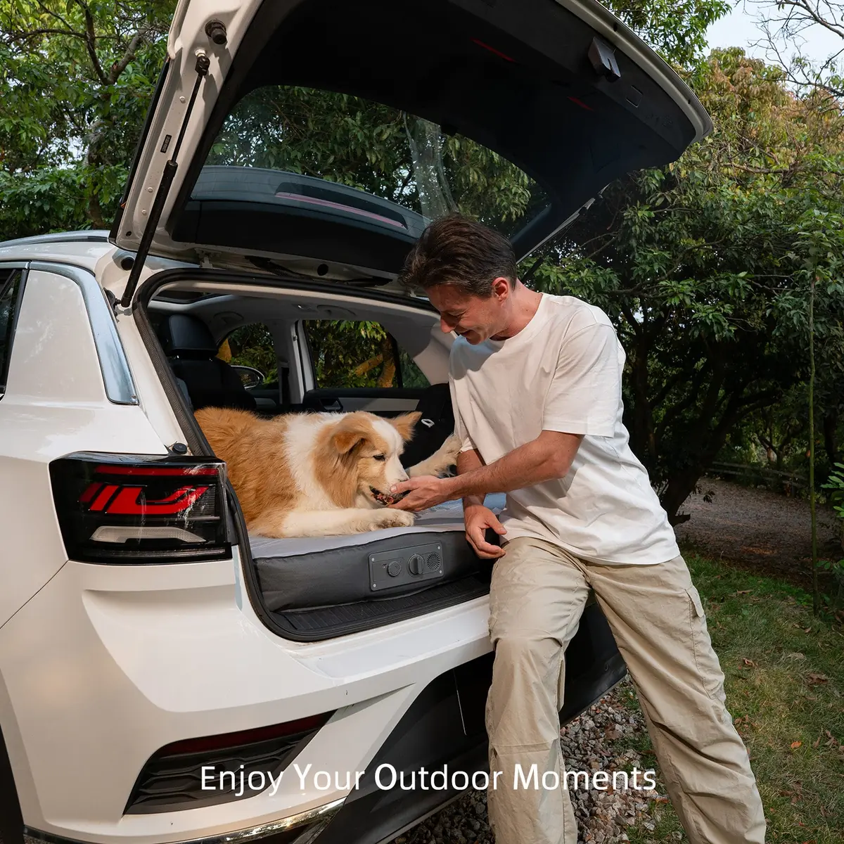 Man feeding a dog a treat at the open tailgate of a white SUV with the mattress visible in the cargo area